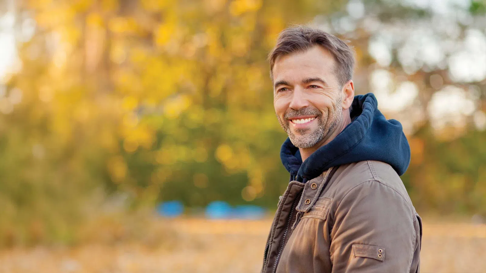 Smiling middle-aged man wearing a brown jacket and blue hoodie in an autumn park with blurred yellow foliage.