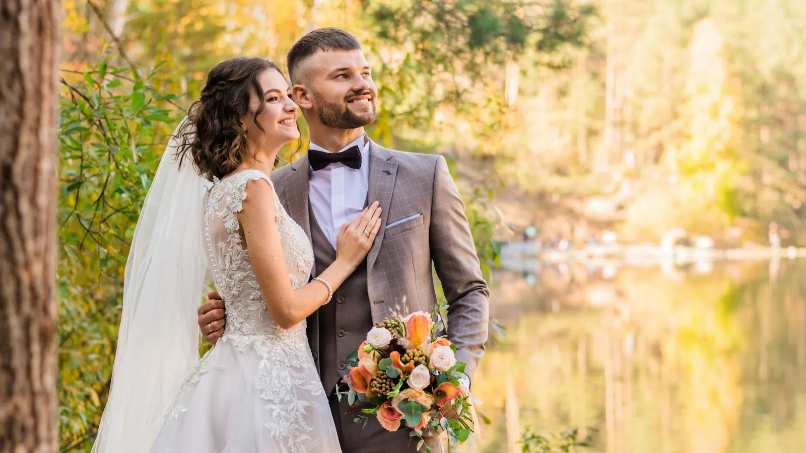 Happy bride and groom in wedding attire standing by a lake surrounded by autumn trees during golden hour
