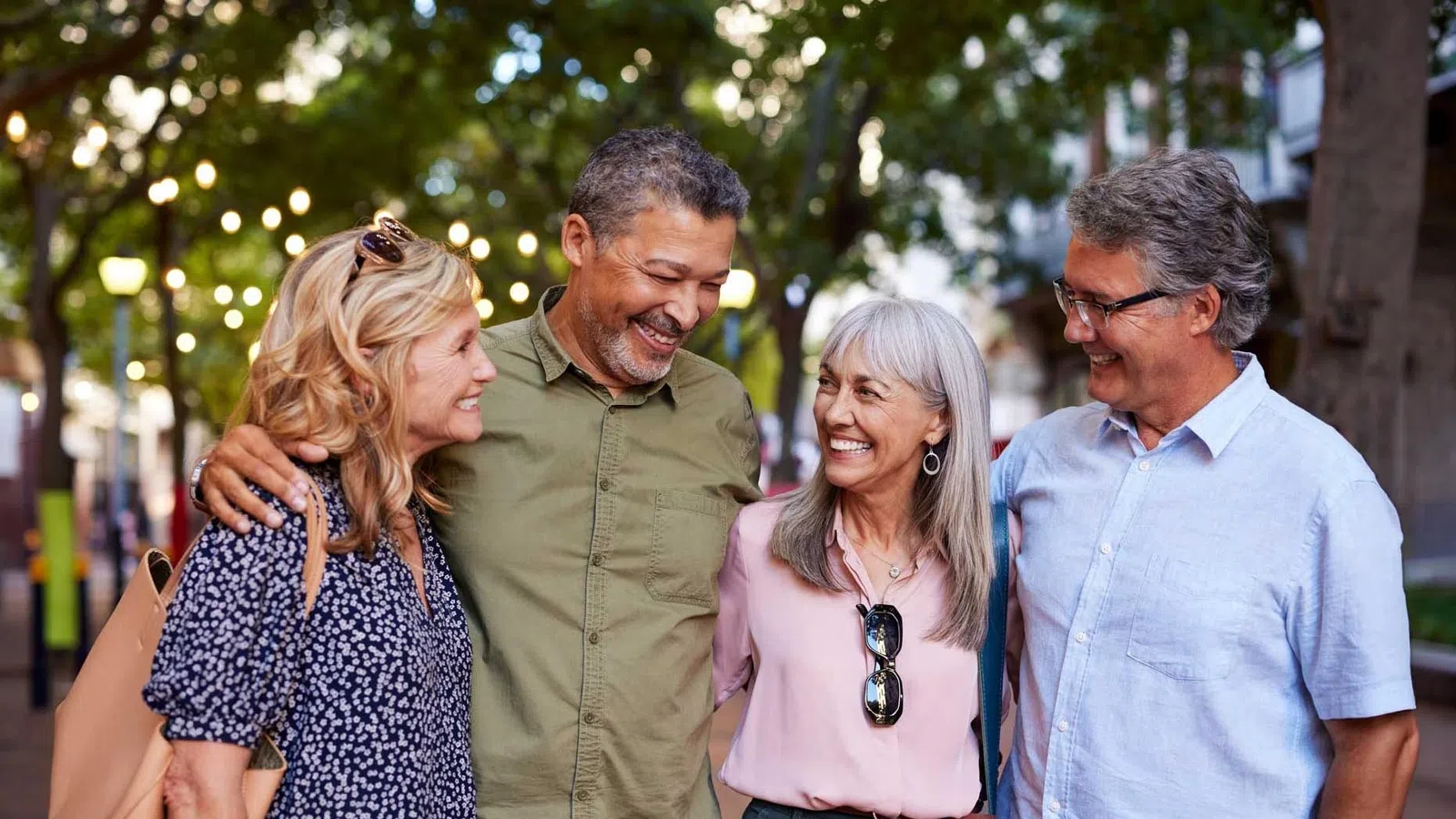 Four middle-aged friends smiling and walking together outdoors under string lights and green trees in the evening.