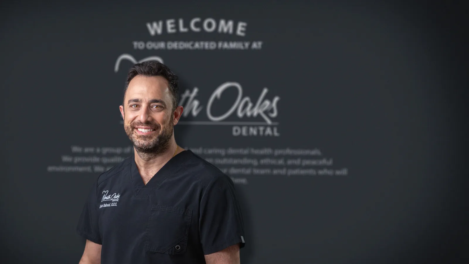 Smiling male dentist in black scrubs stands in front of North Oaks Dental welcome sign on dark wall.