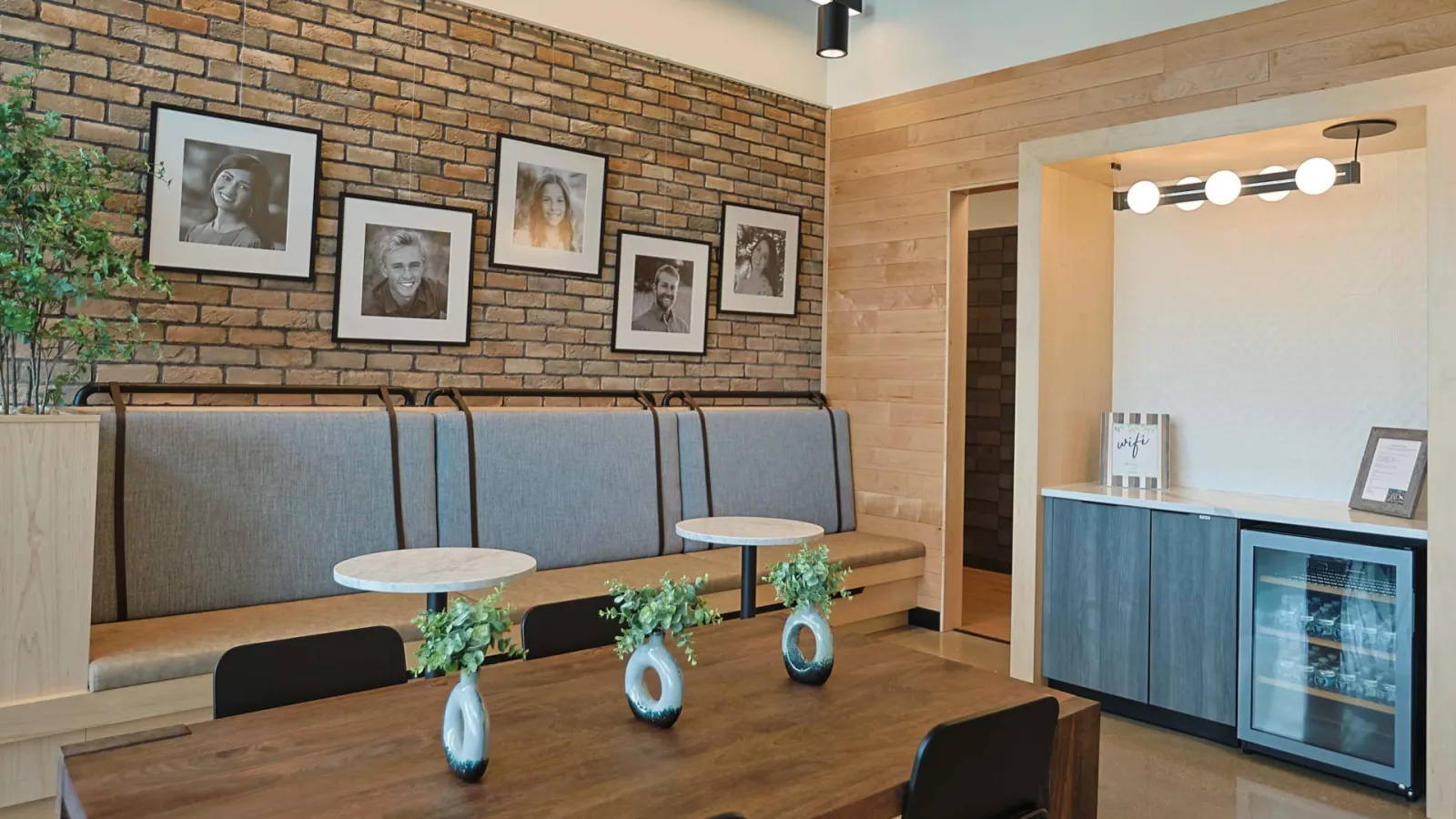 Modern dining area with wooden table, black chairs, framed portraits, brick wall, and built-in bench seating.