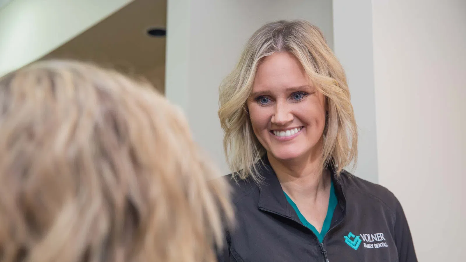 Smiling woman with blonde hair looking in a mirror wearing a black jacket with a dental clinic logo