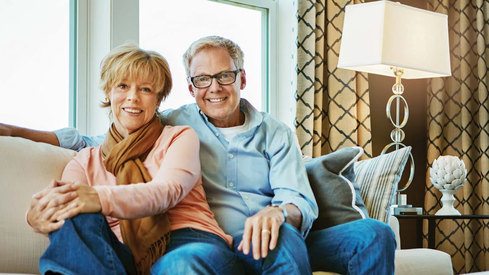 Smiling middle-aged couple sitting closely on a beige sofa in a bright living room with patterned curtains