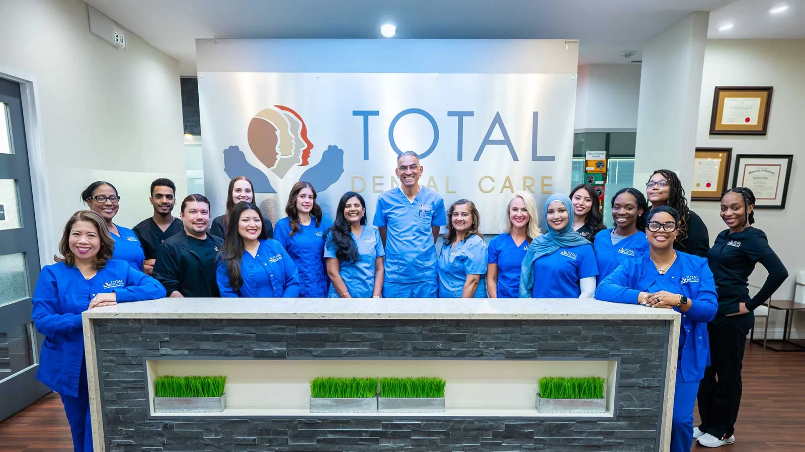 Dental care team standing behind reception desk inside Total Dental Care clinic with logo wall behind.