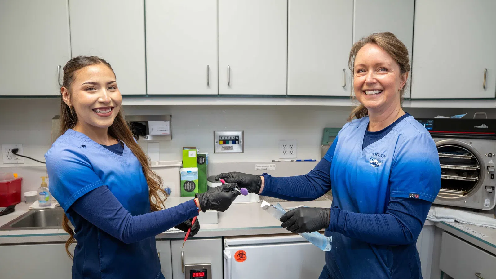 Two female lab technicians in blue scrubs smiling and handling a test tube in a clinical lab setting.