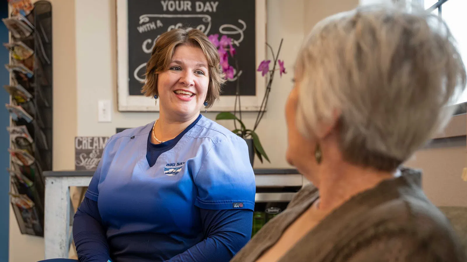 Smiling nurse in blue scrubs talking to elderly woman in a cozy indoor setting with plants and decor.