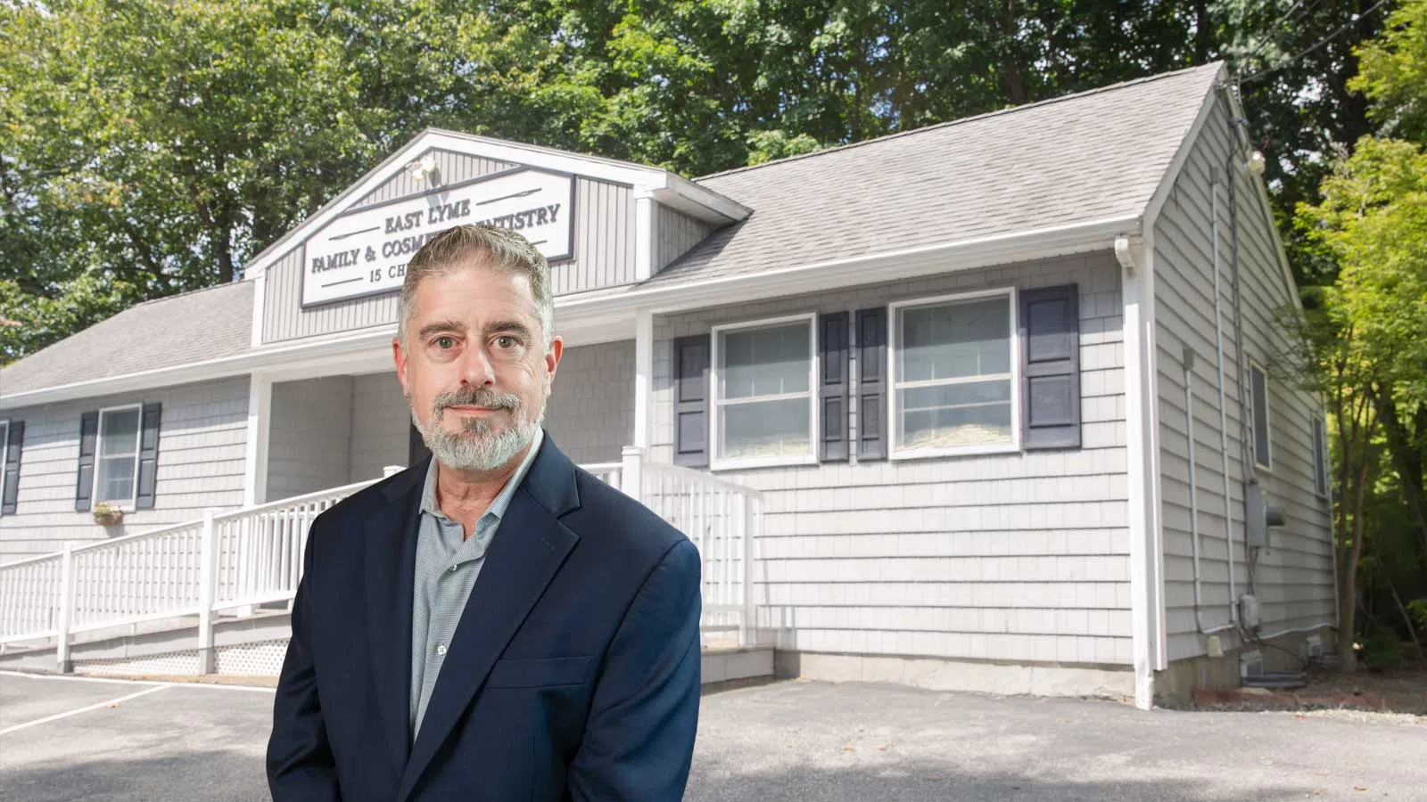 A man in a suit stands outside East Lyme Family & Cosmetic Dentistry building with trees in the background.