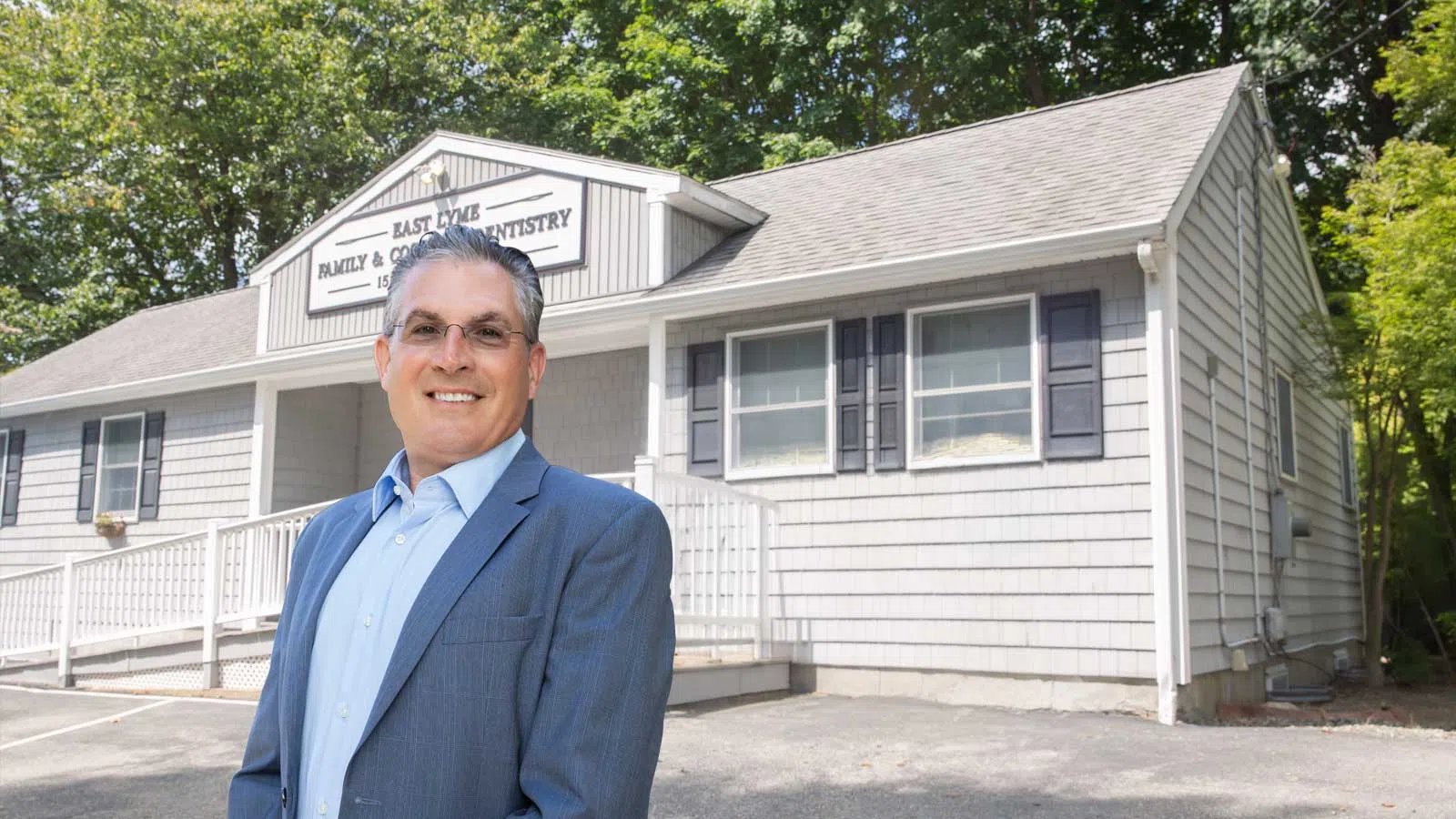 Smiling man in suit standing outside East Lyme Family & Cosmetic Dentistry building with trees in background
