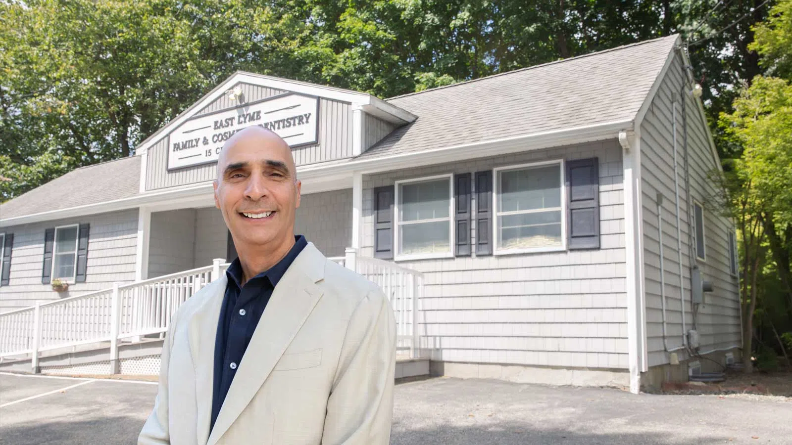 Smiling man in beige blazer standing in front of East Lyme Family and Cosmetic Dentistry building on a sunny day
