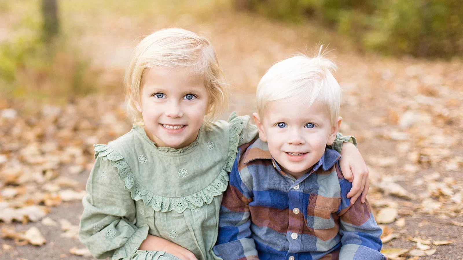 Two young children, a girl in a green dress and a boy in a plaid shirt, sitting on a leaf-strewn path smiling.