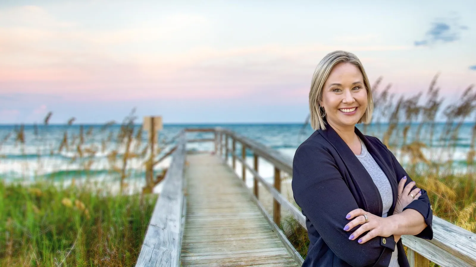 Smiling woman with crossed arms standing on wooden boardwalk by the ocean during sunset.