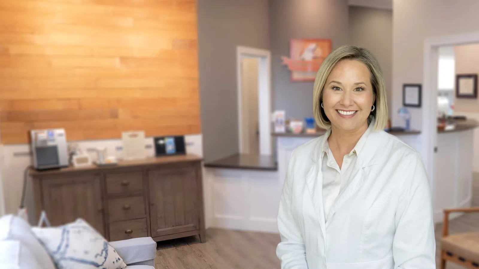 Smiling female doctor in white coat standing in a modern medical office waiting room with wood and neutral decor.