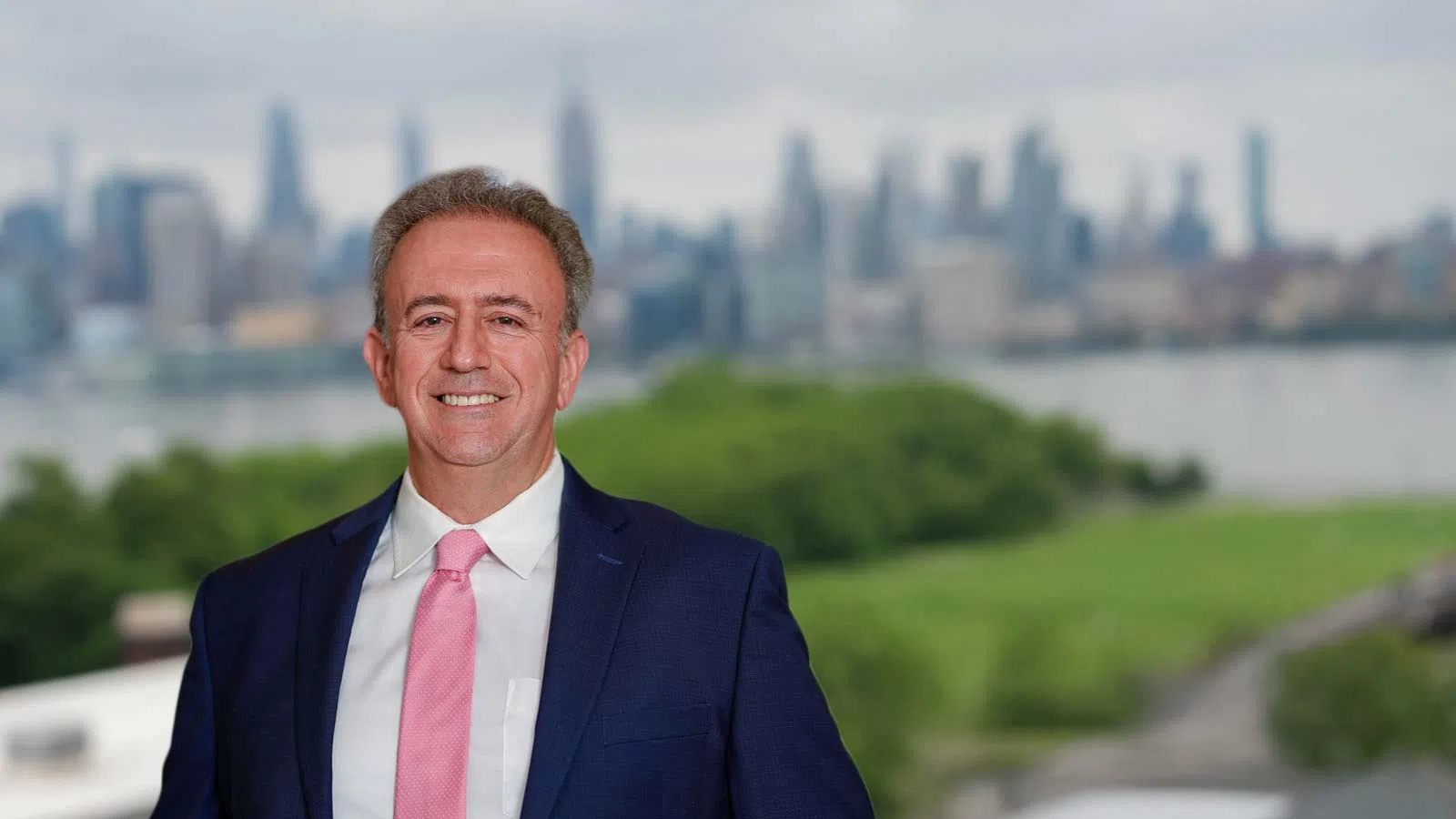 Man in a navy suit and pink tie smiling with a blurred city skyline and greenery in the background