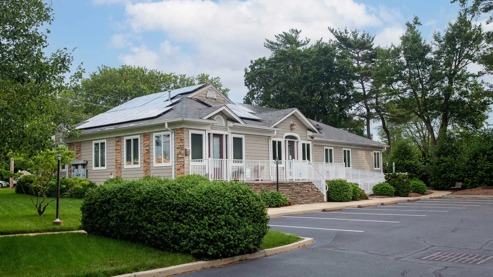 Single-story house with solar panels on the roof surrounded by greenery and an empty parking lot under a blue sky.