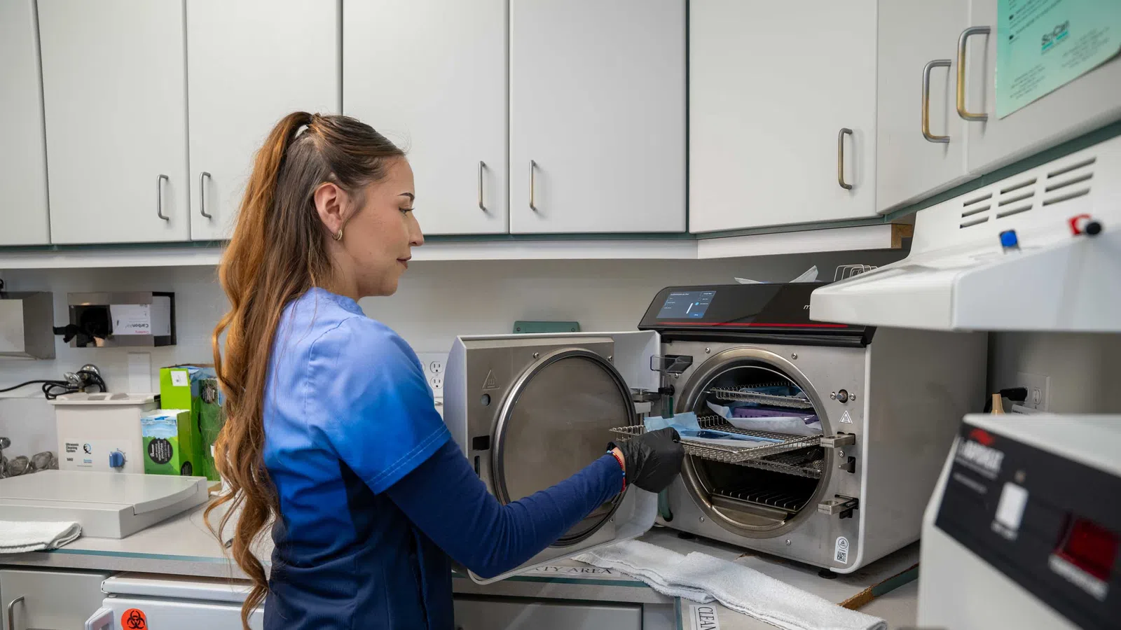Dental professional in blue scrubs operating autoclave sterilizer in a clean, organized dental clinic workspace.