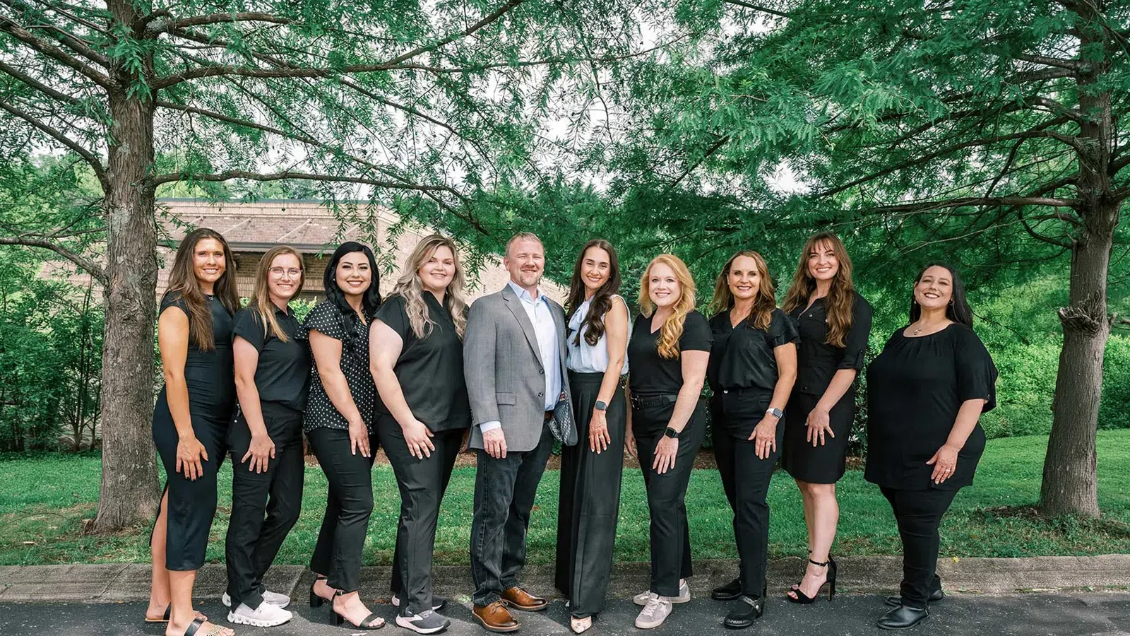 Group photo of nine women and one man standing outdoors in business casual attire under trees on a sunny day.