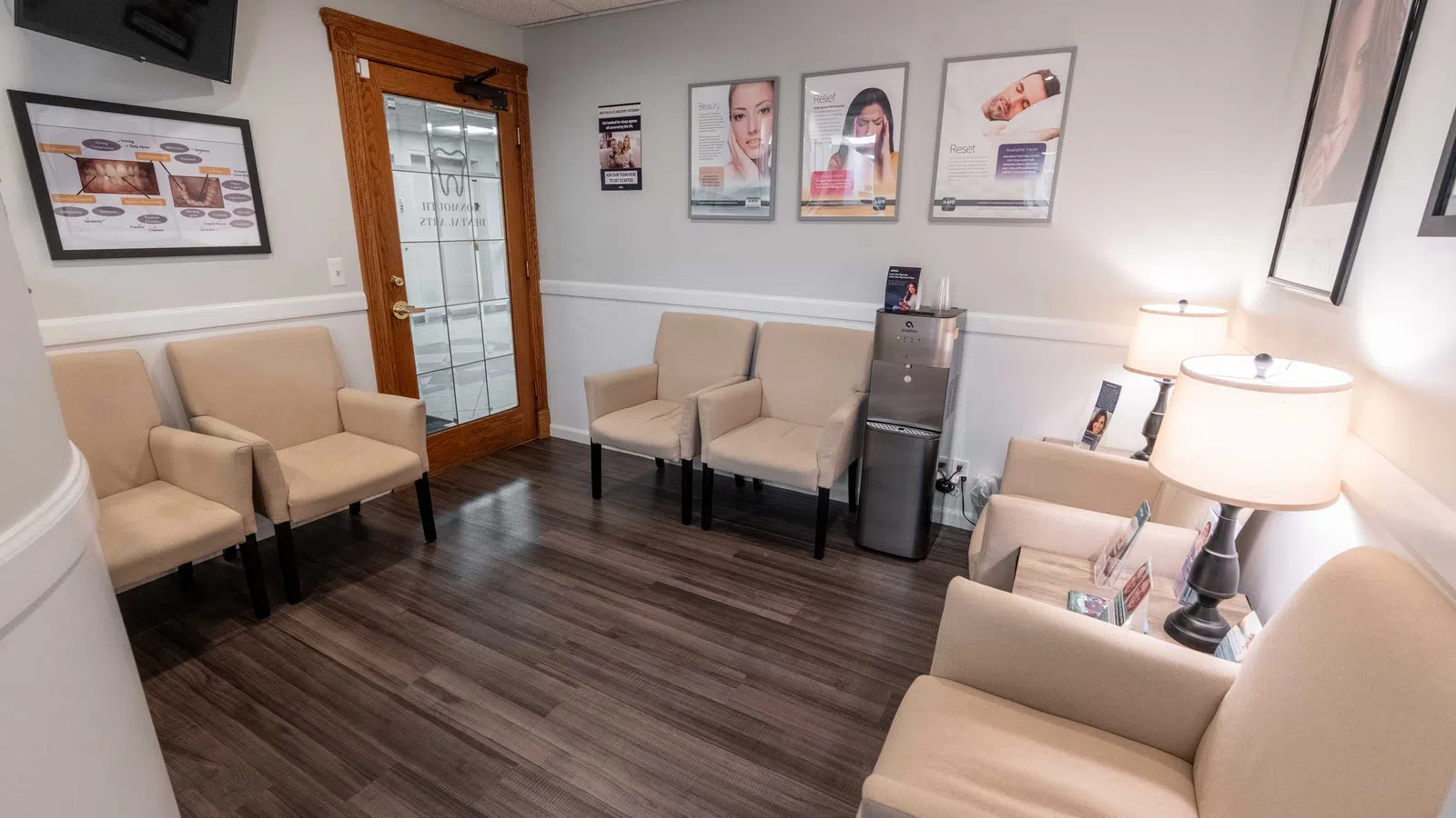 Bright waiting room with beige chairs, wooden floors, skincare posters, and a water cooler near a glass door.