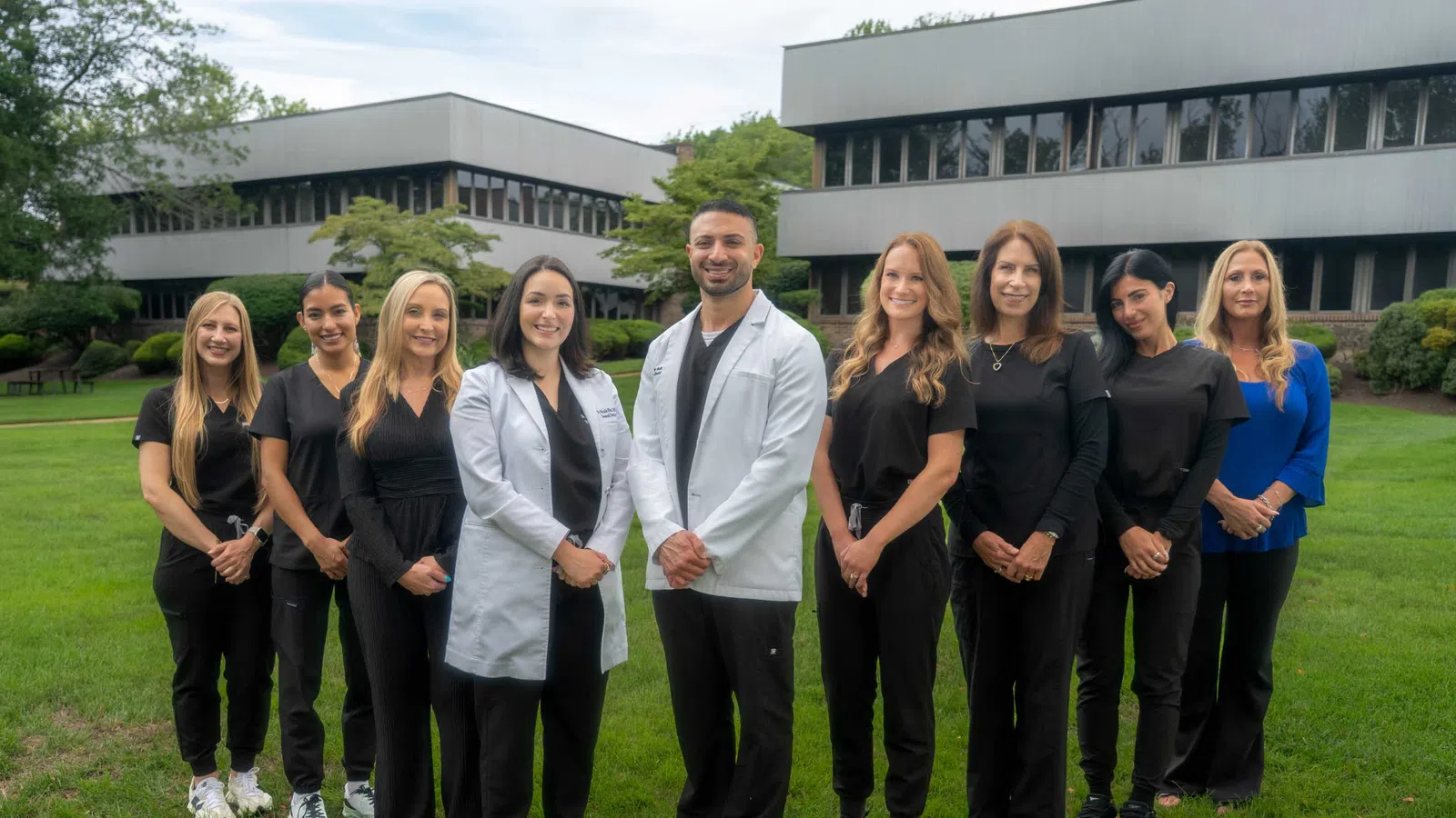 Group of healthcare professionals posing outdoors in front of office buildings, smiling and wearing medical attire.
