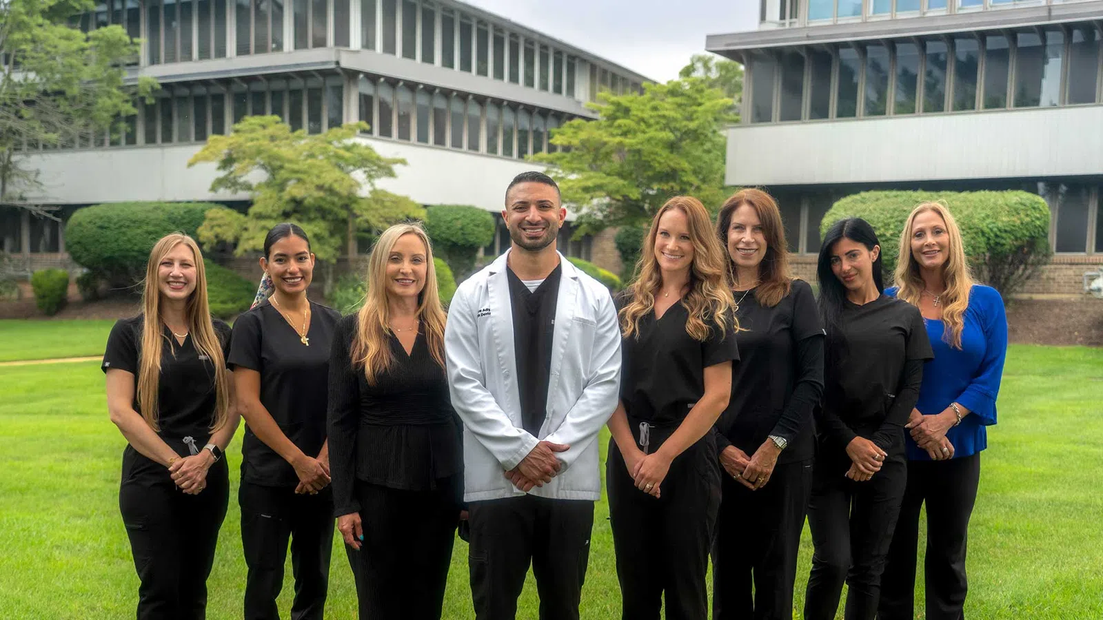Group of eight diverse healthcare professionals standing outdoors in front of office buildings on green lawn