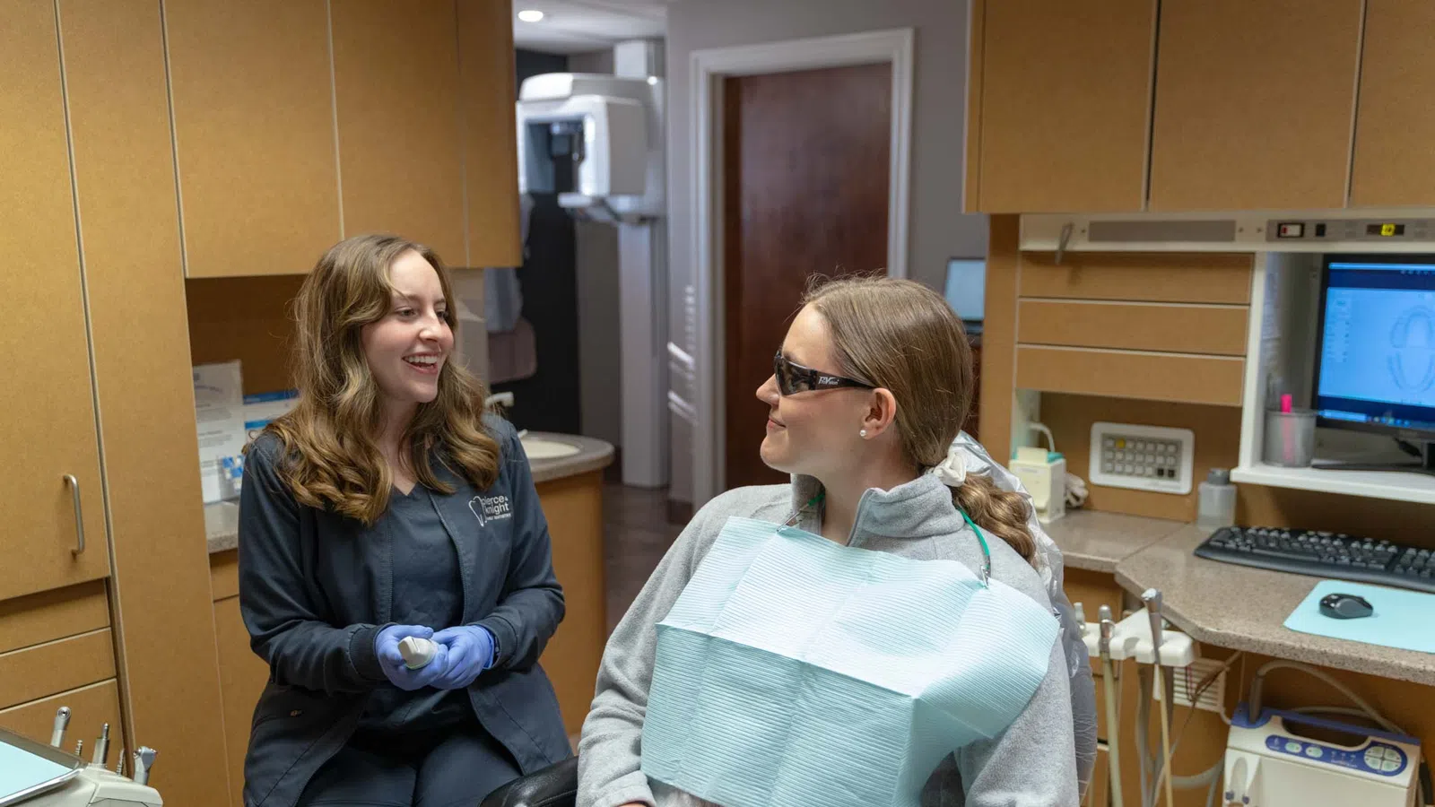 Dental hygienist in gloves talking to patient in dental chair wearing protective glasses and bib in clinic.