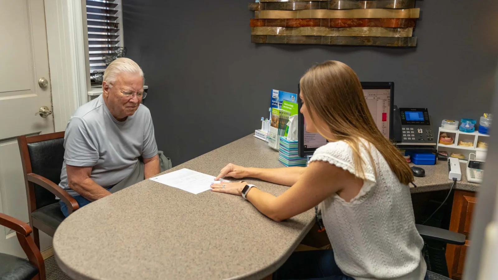 Older man consulting with a female professional at a desk in an office setting with documents and computer.