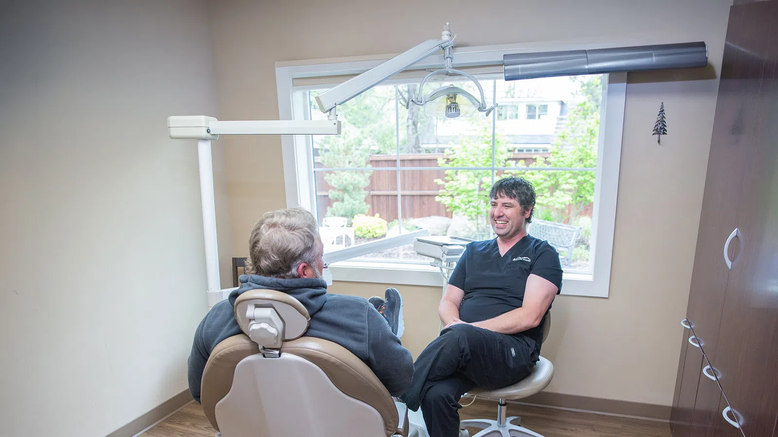 Dentist and patient having a friendly conversation in a bright dental office with a large window and equipment.