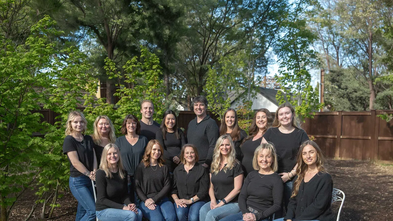 Group of fifteen diverse adults posing outdoors in casual black tops and jeans with trees and wooden fence background