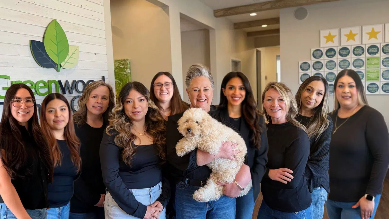 Group of smiling women wearing black tops posing indoors with a fluffy white dog in front of a Greenwood sign.
