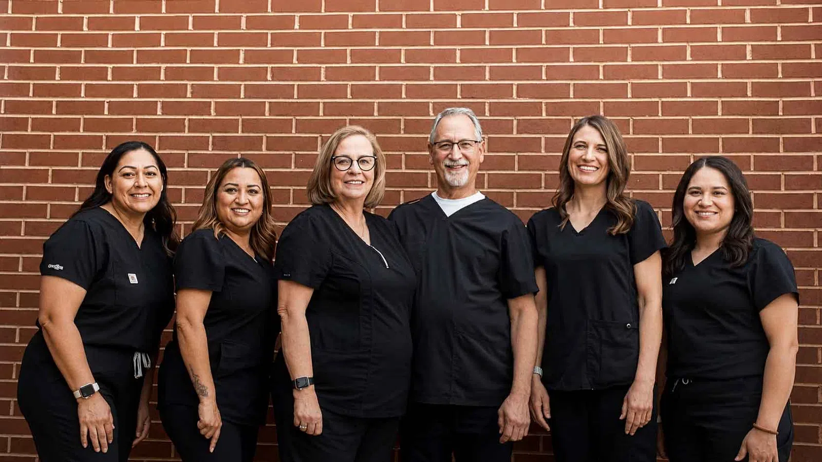 Group photo of six smiling healthcare professionals wearing black scrubs standing against a red brick wall.