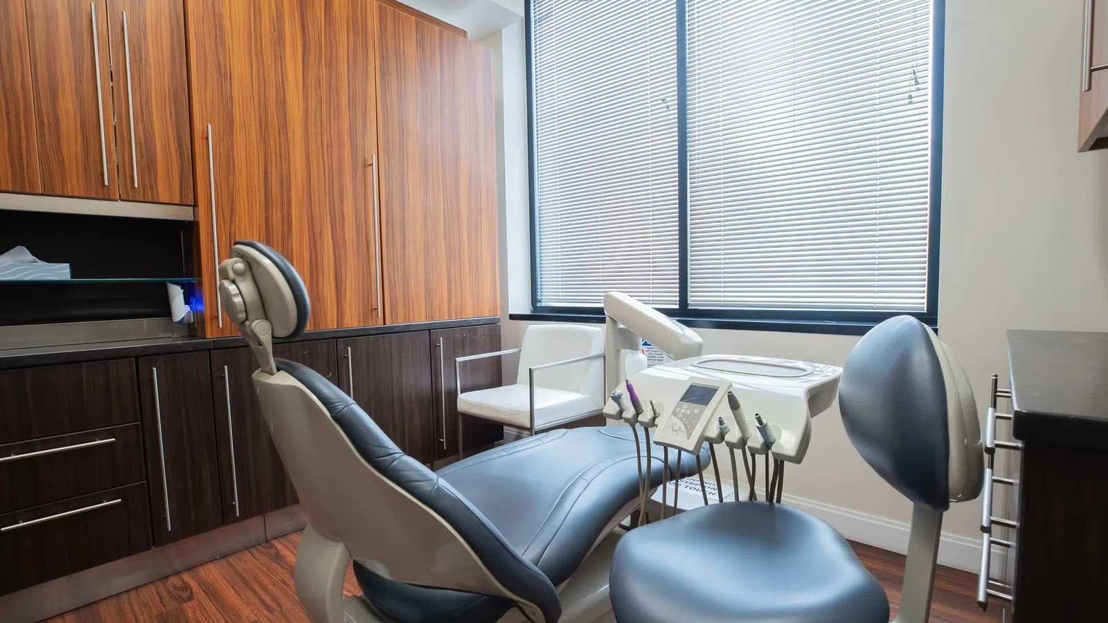 Modern dental clinic room with dental chair, equipment, wooden cabinets, and large window with blinds.