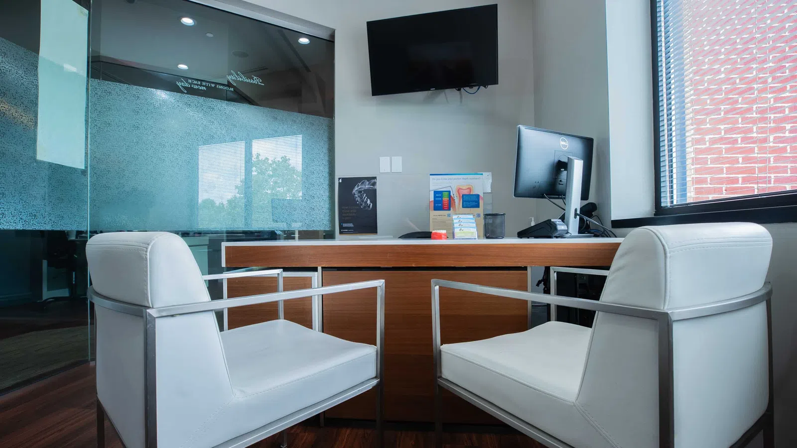 Modern office with two white chairs facing a wooden desk, computer monitor, TV, and large window with blinds.