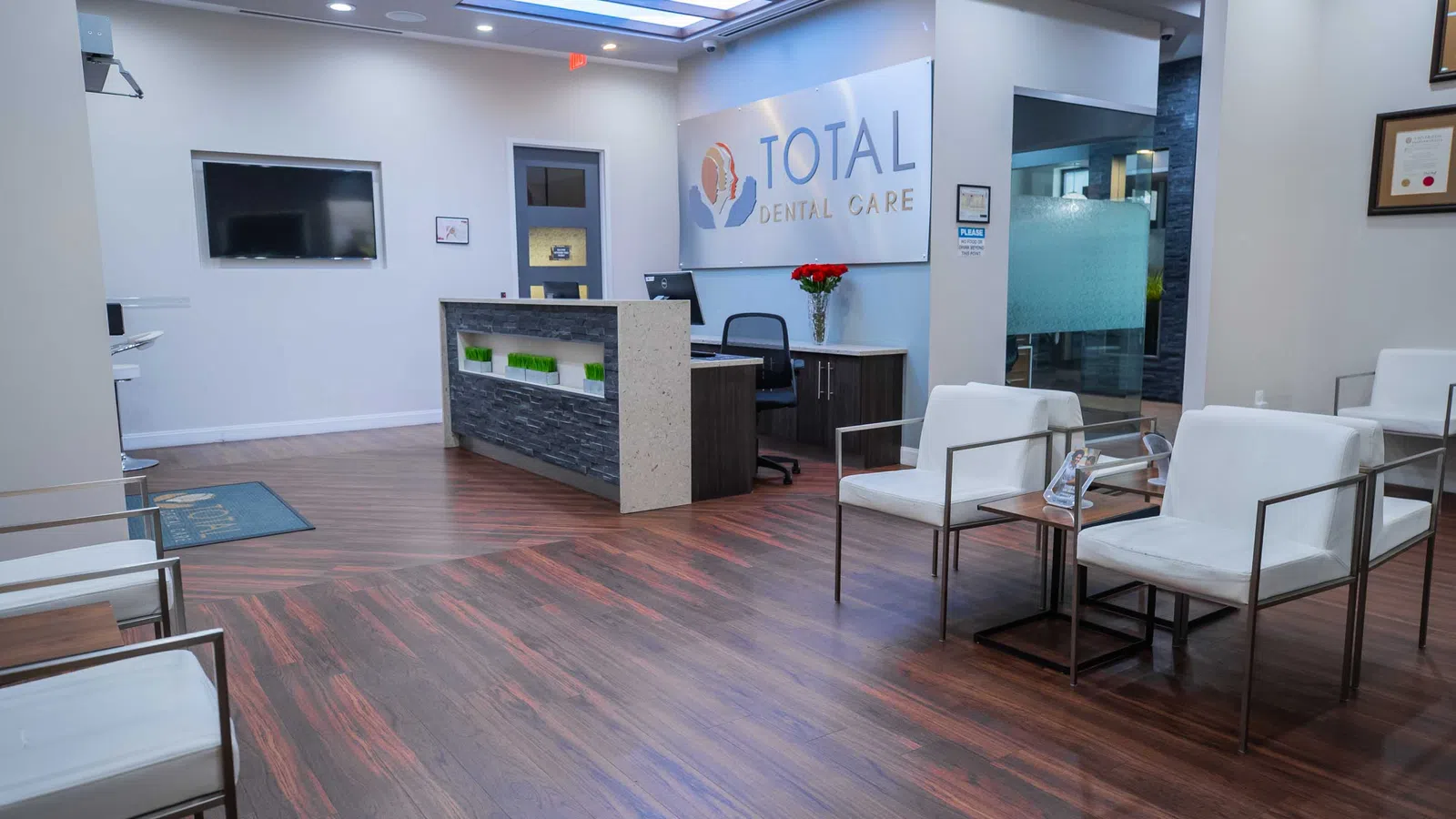 Modern dental office waiting area with white chairs, wood flooring, reception desk, and Total Dental Care sign.
