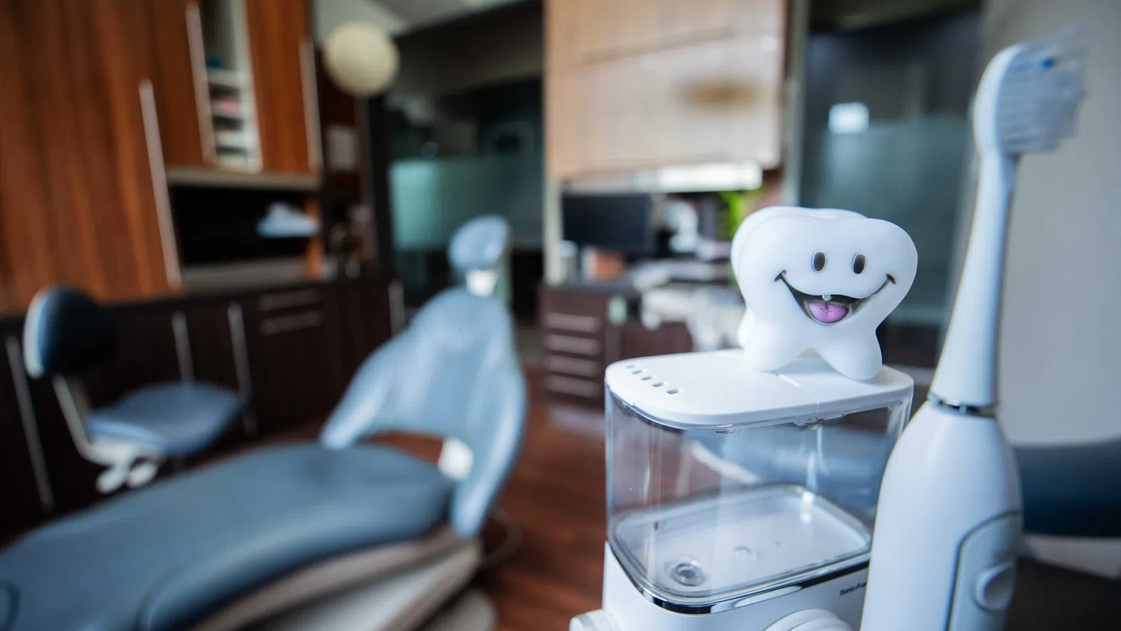 Dental clinic setup with electric toothbrush, water flosser, and smiling tooth model in foreground, dental chair blurred.