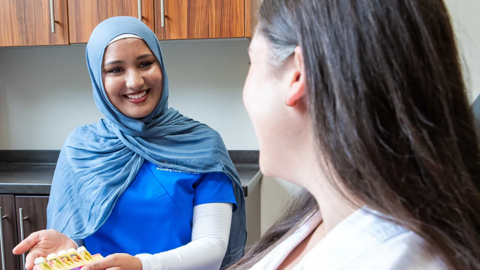 Smiling female dental professional in blue scrubs and hijab explaining teeth model to patient.