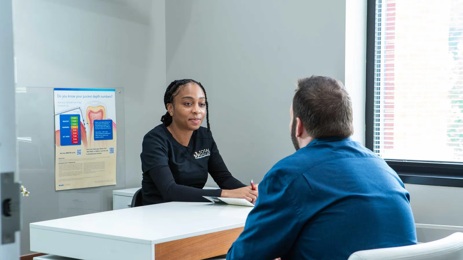 Female healthcare professional consulting a male patient in a modern office with dental health poster visible.