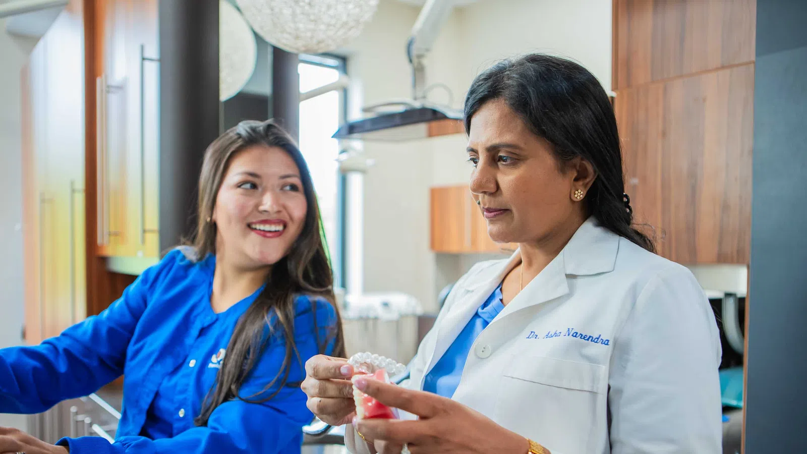 Dentist in white coat showing dental model to smiling assistant in blue scrubs at clinic.