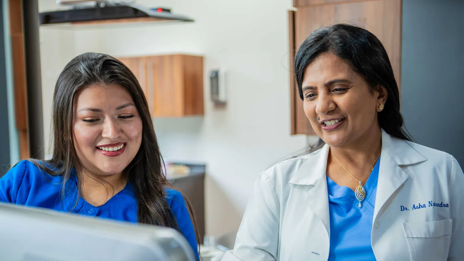 Two female healthcare professionals in blue scrubs reviewing information on a computer screen in a medical office.