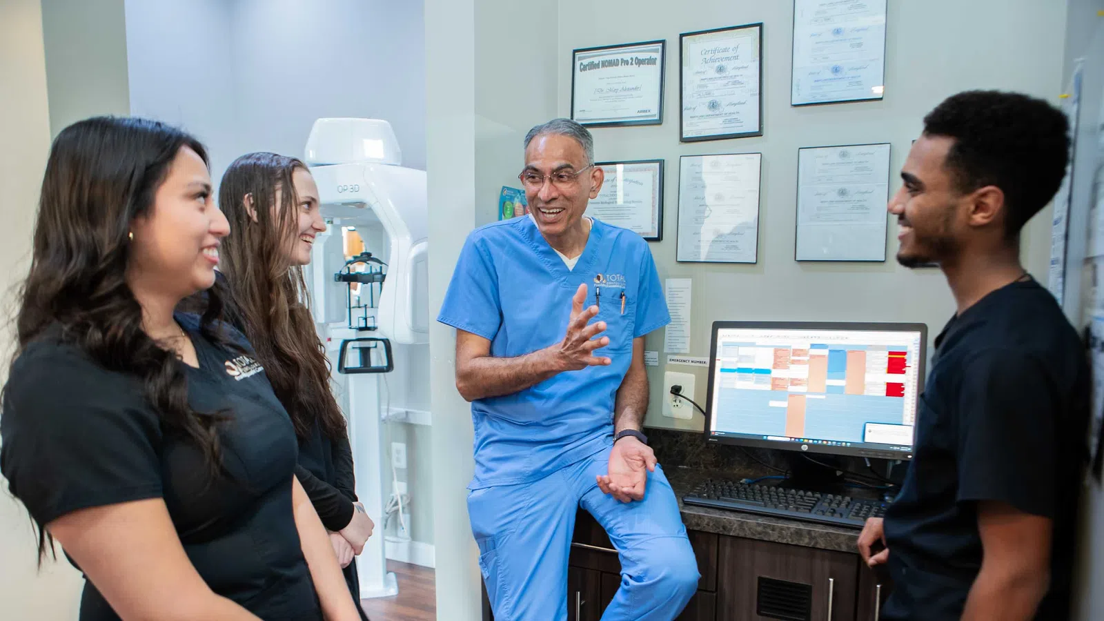 Smiling healthcare professional in blue scrubs talks with three colleagues in a medical office with certificates and computer.