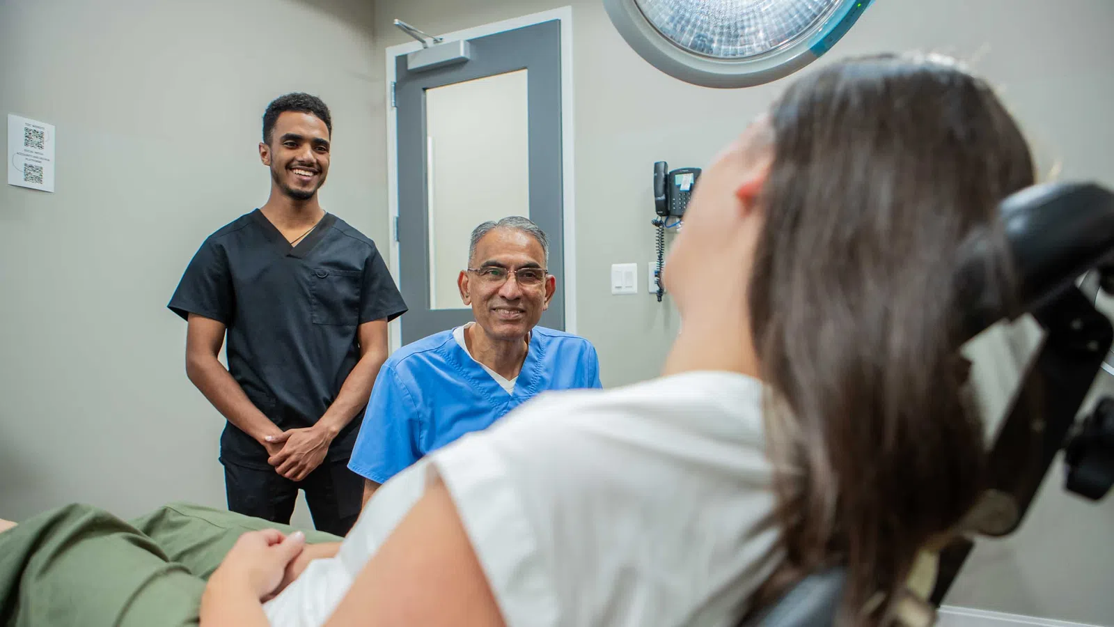 Two healthcare professionals smiling at a patient reclining in a medical examination chair in a clinic room.