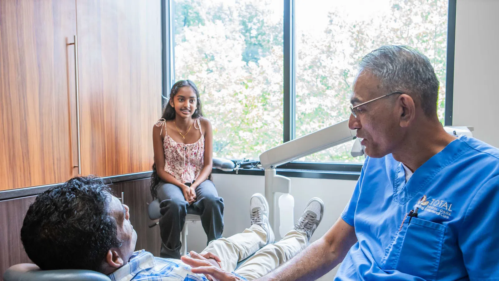 Dentist in blue scrubs talking to male patient in dental chair while young girl sits nearby by window.