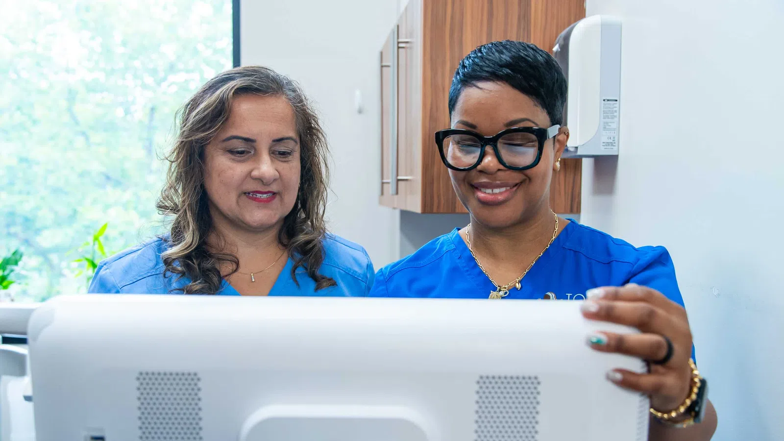 Two female nurses in blue scrubs smiling and looking at a computer screen in a medical office.