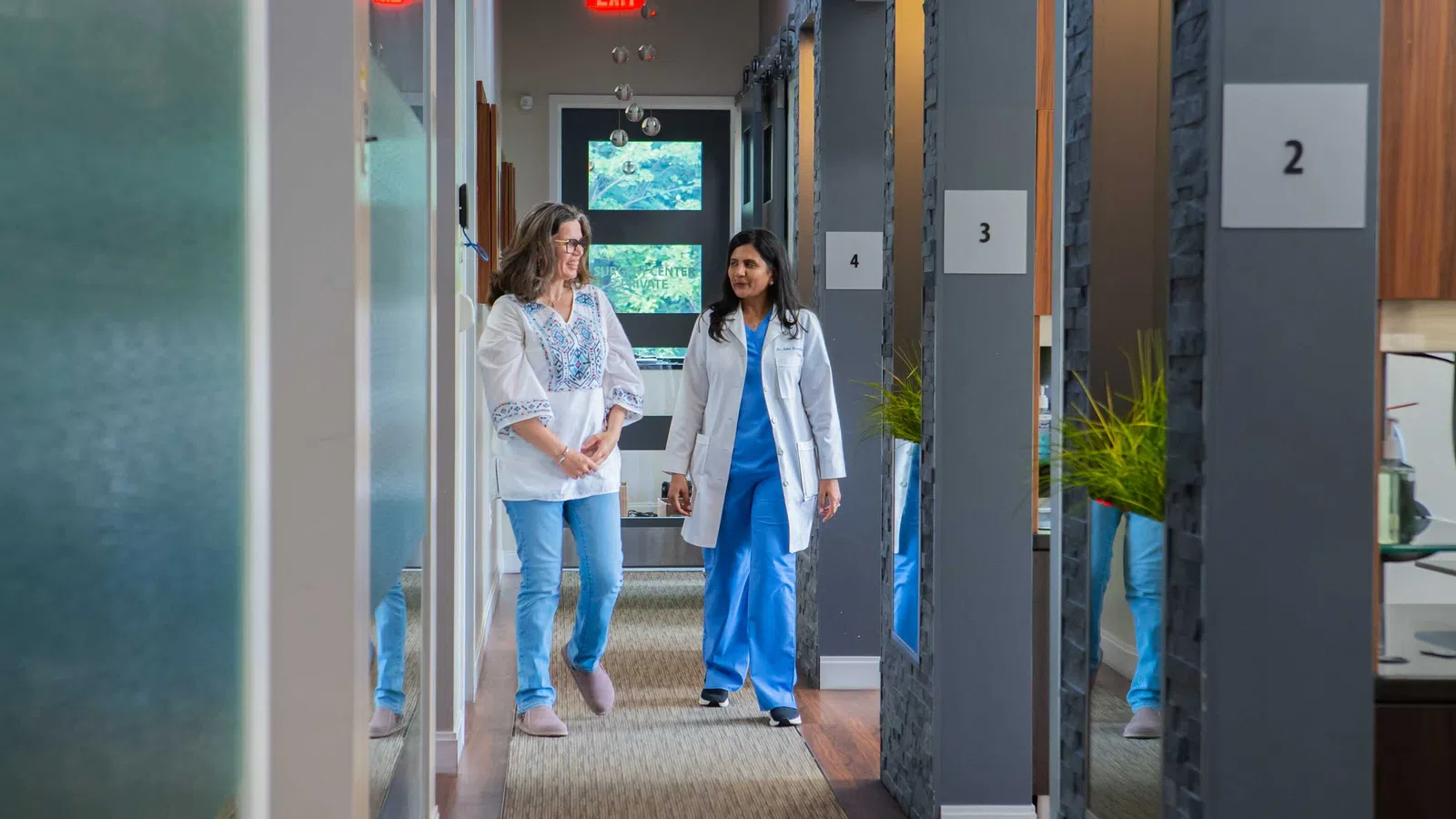 Doctor and patient walking and talking together in a modern medical clinic hallway with numbered rooms.