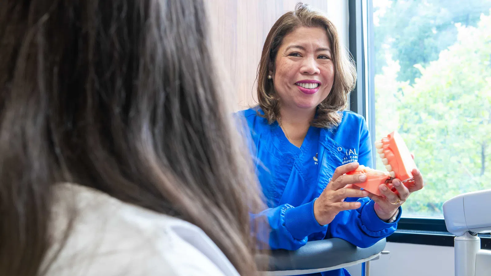 Female dentist in blue scrubs explaining dental care using a tooth model to a patient in a clinic.