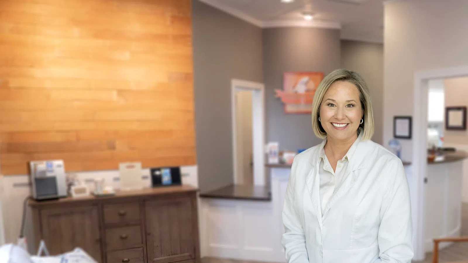 Smiling female healthcare professional in white coat standing in a modern medical office reception area.