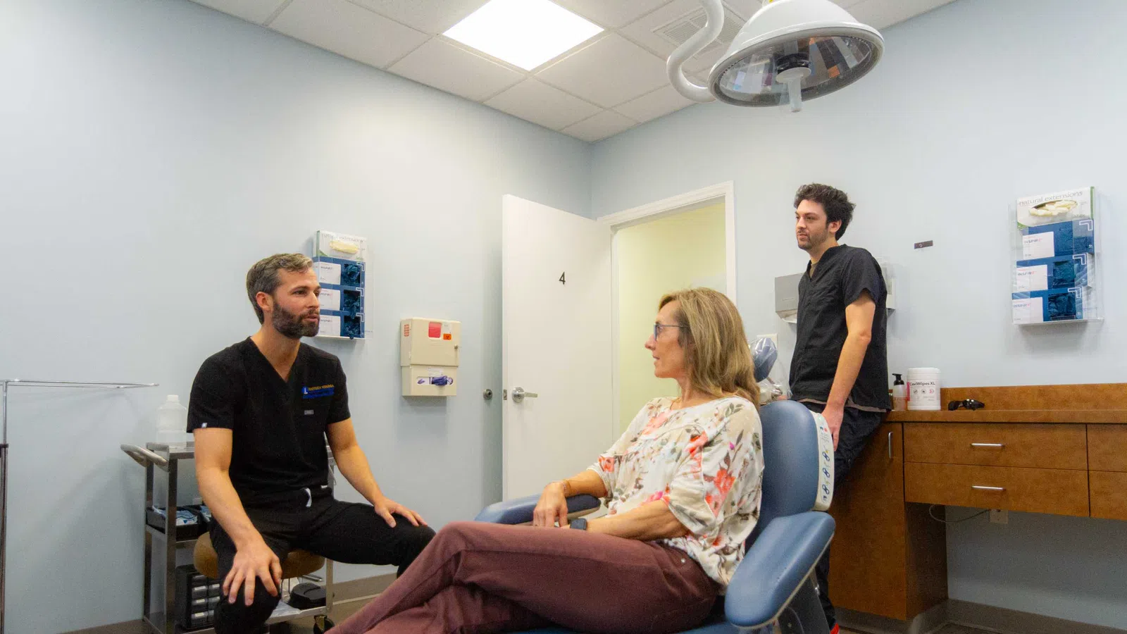 Patient sitting on dental chair talking with two male dentists in a clean, well-lit clinic room