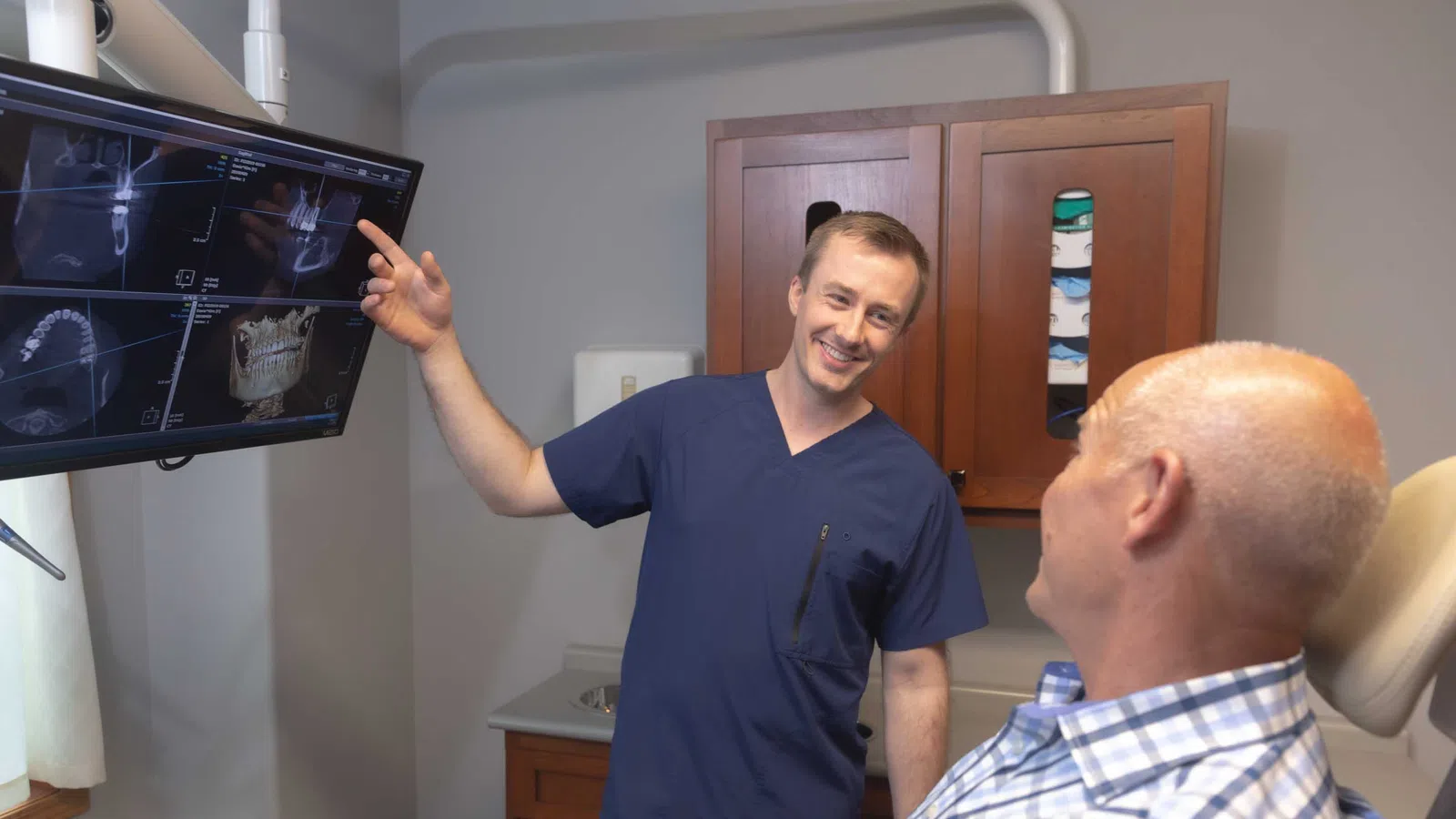 Dentist in blue scrubs showing dental X-rays on screen to an older male patient in a dental office.