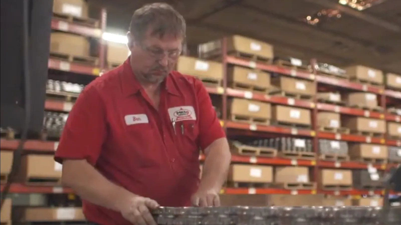 Worker in red shirt handling metal parts on a conveyor in a warehouse with shelves full of boxes.