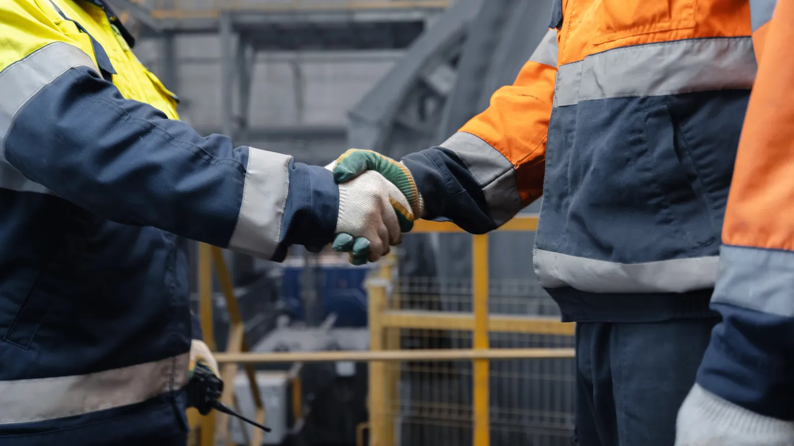 Two industrial workers wearing safety jackets and gloves shaking hands inside a factory setting.