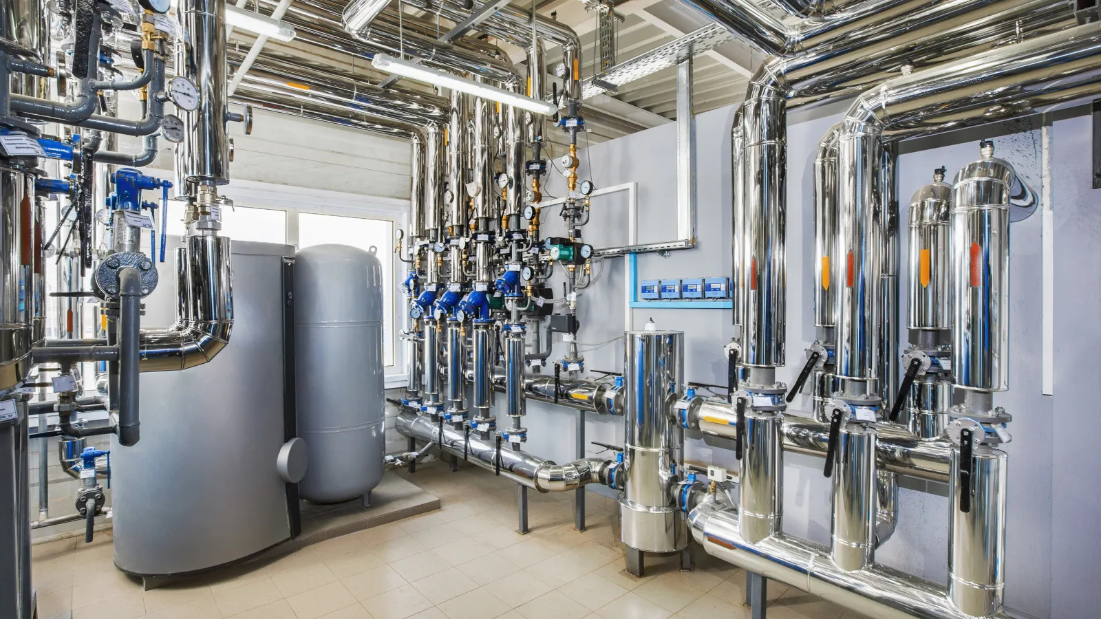 Industrial mechanical room with insulated pipes, pressure gauges, valves, and large tanks for fluid handling.