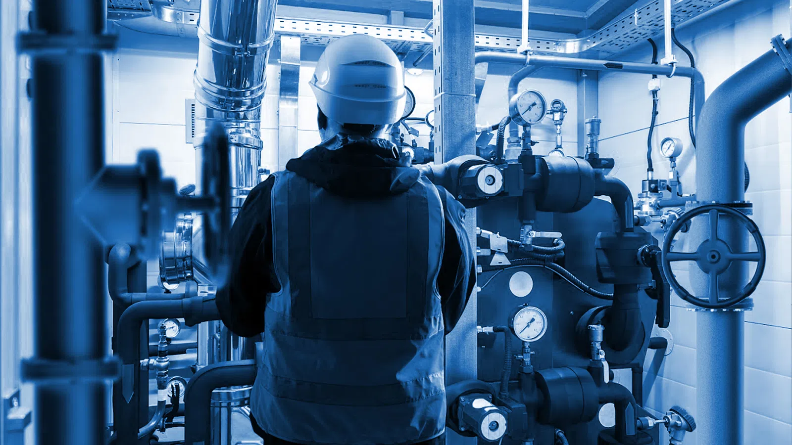Engineer in safety gear inspecting complex industrial piping and gauges in a mechanical room with blue lighting.